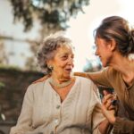 Una persona adulta tiene la mano sulla spalla a una signora anziana, si guardano sorridendo.